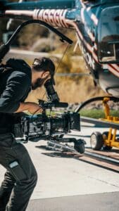 Man in black cap filming with a professional camera near a helicopter on a sunny day